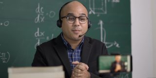 印度年轻教师男子戴着耳机坐着通过智能手机进行在线视频会议直播。亚洲数学教师在线授课，供学生在线学习。