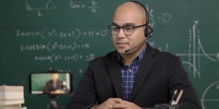 印度年轻教师男子戴着耳机坐着通过智能手机进行在线视频会议直播。亚洲数学教师在线授课，供学生在线学习。
