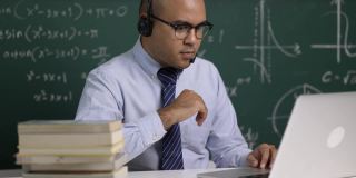 印度年轻教师男子戴着耳机坐着用笔记本电脑进行在线视频会议直播。亚洲数学教师在线授课，供学生在线学习。