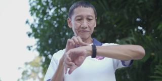 亚洲老年男子在公园的树下触摸智能手表活动跟踪器。手指敲击运动手表卡路里和心率信息。慢动作轨道拍摄。卫生保健的概念。