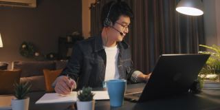 一名亚洲男子在家里工作到很晚，在视频电话会议中使用笔记本电脑，并在笔记本上写字
