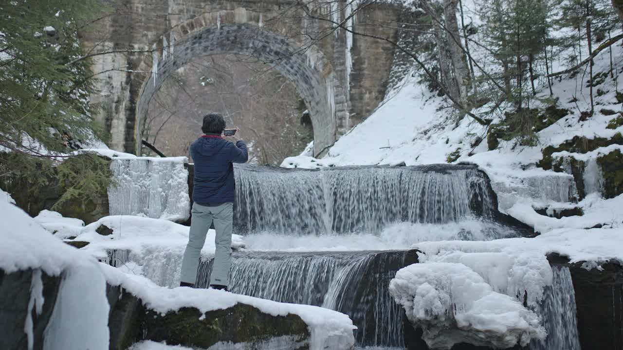 独自一人在自然。一名男性游客在冬季拍摄美丽的冰冻瀑布。旅行和摄影。