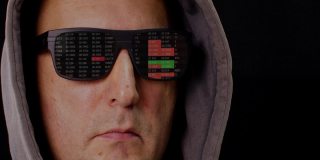近距离观察蒙面男子和黑色VR眼镜与反射交易交易数据。证券交易所里映着黑色墨镜的男子戴着兜帽，孤立在黑暗的背景下