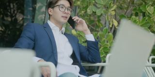 年轻聪明迷人的亚洲自信男性穿着正装，布，手持智能手机，在写字楼庭院的现代花园与客户沟通，随意咨询