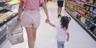 年轻的亚洲美丽的母亲拿着购物篮和她的孩子走在超市。她正在为她的小孩挑选架子上的食物。健康购物在零售商店。