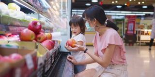 年轻美丽的亚洲母亲提着购物篮和她的孩子在超市行走，戴着口罩，以预防新冠肺炎感染。她正在挑选红苹果水果并从货架上挑选。