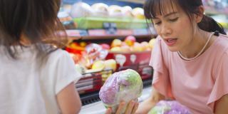 年轻的亚洲美丽的母亲拿着购物篮和她的孩子走在超市。她从货架上挑选绿色沙拉蔬菜，然后给她的小孩吃。健康的购物。
