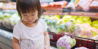 年轻的亚洲美丽的母亲拿着购物篮和她的孩子走在超市。她从货架上挑选绿色沙拉蔬菜，然后给她的小孩吃。健康的购物。