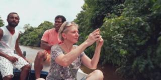 斯里兰卡本托塔河上，一名留着短发的白人老年妇女，在两名当地导游的陪同下，在一艘船上用她的智能手机拍照。