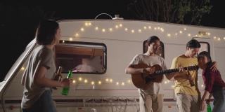 一群亚洲男女朋友在晚上的户外聚会上一起弹吉他。朋友们带着幸福和微笑碰杯喝啤酒。乘露营车旅行大自然。