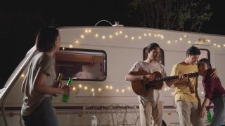 一群亚洲男女朋友在晚上的户外聚会上一起弹吉他。朋友们带着幸福和微笑碰杯喝啤酒。乘露营车旅行大自然。视频素材模板下载