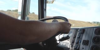 在乡村公路上驾驶卡车时，男性卡车司机的手握着方向盘。卡车司机开车穿过乡村前往目的地。物流和运输的概念。近距离