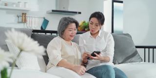 亚洲家庭妇女和她的母亲使用智能手机教玩互联网一起在客厅在家里的幸福和微笑。两代沟技术使用生活方式的概念。
