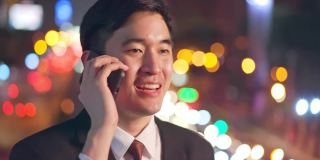 亚洲年轻商人站在户外使用手机在城市的市中心晚上。他在用智能手机聊天，笑得很开心。这位商人对现代网络技术很满意。