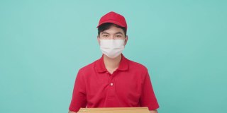 孤立背景，身穿红色制服的亚洲快递员戴着面具，手里拿着一个中等大小的包裹，在前面让步。男邮差竖起大拇指，面带微笑。交付的概念