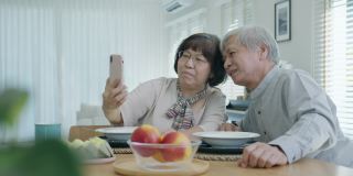 年轻的老年亚洲公民夫妇手持手机自拍视频在线通话在家中公寓的餐桌上老年人用数字技术虚拟谈话会议的概念。