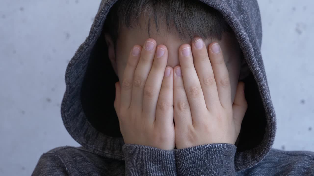 悲伤沮丧的孤独男孩在兜帽夹克用手掩盖他的脸。白天照明，灰色墙壁背景