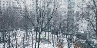 冬天冰雪覆盖的城市公园。在光秃秃的树木上伸展树枝。雪花正在慢慢地飘落。操场上有滑梯、长椅、单杠、停车带车。城市生活