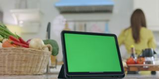 绿色屏幕的平板电脑，美丽的背景，在家里做饭，快乐的脸，妻子在她的家快乐地做早餐。
