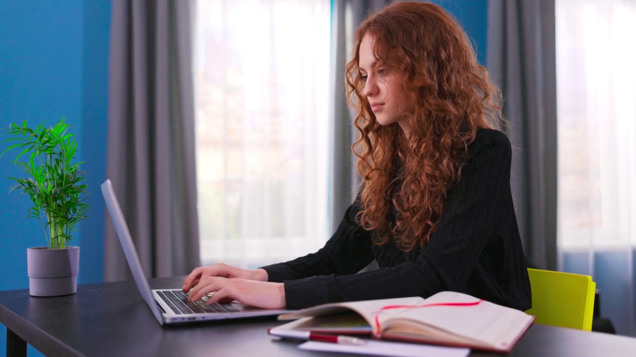 美丽的少女坐在一个舒适的客厅的桌子上，并在电脑上跟随一个视频学校课程。远程学习。在社交网络上与朋友聊天