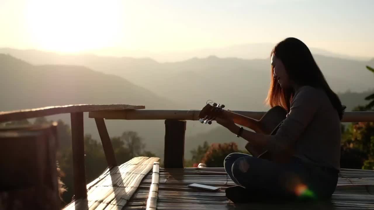 一个女性坐在和弹吉他的慢动作在一个美丽的山景日落前的木制阳台上
