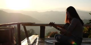 一个女性坐在和弹吉他的慢动作在一个美丽的山景日落前的木制阳台上