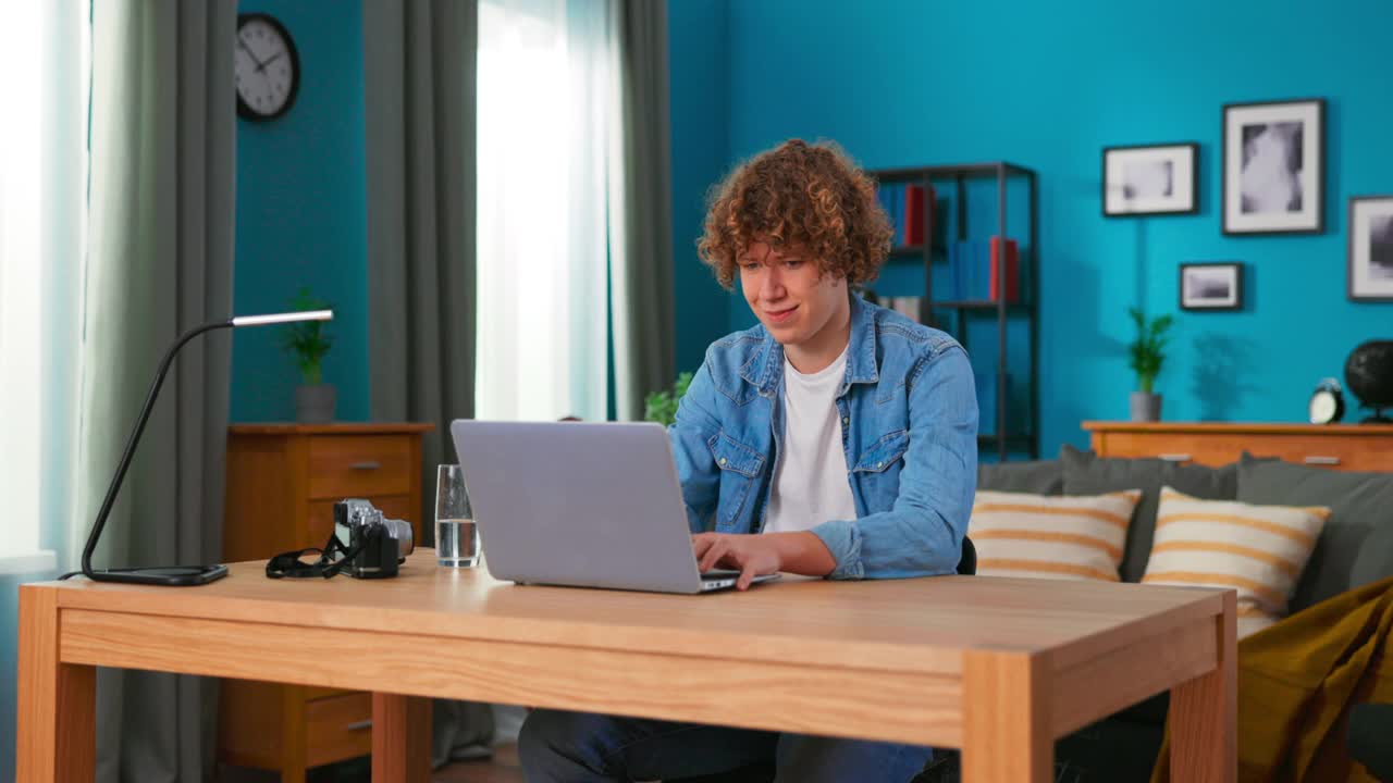 英俊的白人男孩在笔记本电脑上工作，而坐在椅子上在时尚舒适的客厅。自由职业者在家工作。浏览互联网，玩得开心