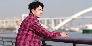 英俊的中国青年坐在黄浦江畔，以大桥和城市为背景，4k慢镜头。
