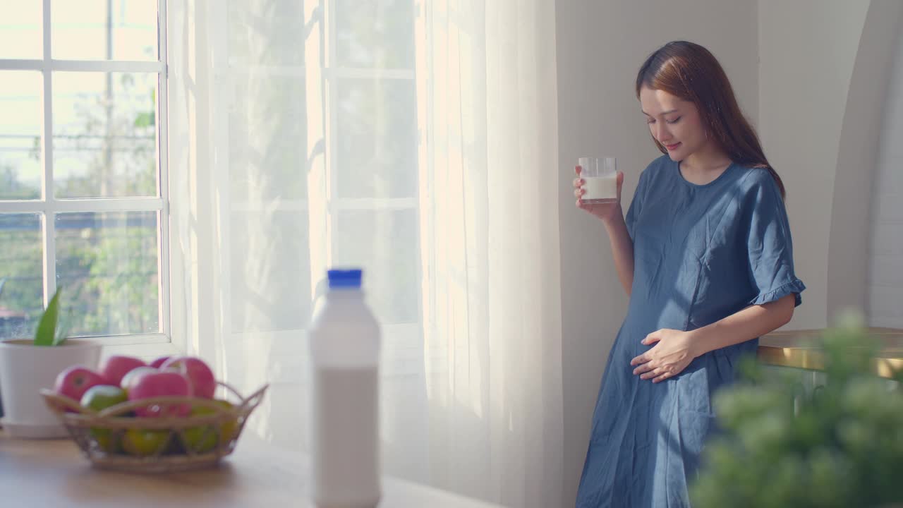 一位孕妇正在喝牛奶，看着窗户，轻轻地摸着她的肚子，怀孕/分娩