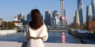 美丽的中国年轻女子转身挥着她的手和上海外滩的地标背景说你好，4k慢镜头。