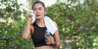 美丽的亚洲健身女人在运动服和毛巾休息后，跑步锻炼喝水。疲惫的女性在户外公园训练后休息