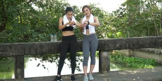 健康的亚洲女性穿着运动服和毛巾，跑步后休息，一起在公园户外用智能手表检查心率和健身进度。两个朋友在锻炼后聊天和休息
