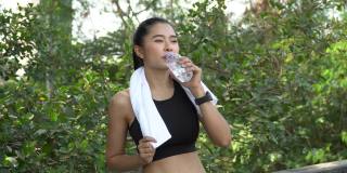 美丽的亚洲健身女人在运动服和毛巾休息后，跑步锻炼喝水。疲惫的女性在户外公园训练后休息