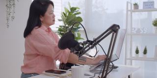 亚洲女性电台主持人录制播客在她的笔记本电脑与耳机和麦克风。女播客制作音频播客从她的家庭工作室的在线节目。