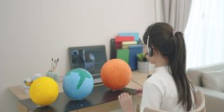 一名亚洲少女通过客厅的笔记本电脑屏幕向老师在线展示她的科学作品，当时学校因COVID-19疫情而停课。