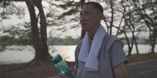 口渴的资深亚洲活跃男子饮用新鲜水从瓶子在树木公园在傍晚日落。成熟的男性在运动时饮用能量饮料来补充疲劳和疲劳的水分。