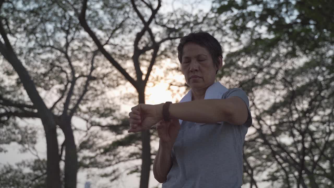 一名亚洲老年妇女在公园的树下触摸智能手表活动跟踪器。手指敲击运动手表卡路里和心率信息。慢动作轨道拍摄。卫生保健的概念。