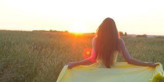 快乐年轻美丽的女人穿着黄色的衣服在麦田里奔跑在日落的夏天，自由健康的幸福理念。美女长着长发，自然在户外，举起双手徐莫太阳镜头耀光