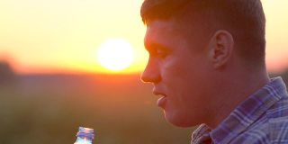 一个年轻人在麦田里喝着从一个塑料瓶里流出的清水。日落时分，有人在户外喝水。夏天。太阳眩光。慢镜头特写