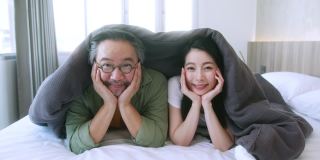 幸福快乐的成熟成人亚洲已婚夫妇家庭的妈妈爸爸躺在卧室的白色软床上笑放松休闲周末活动，俯视图手持拍摄慢动作