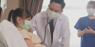 亚洲医生在康复室的床上用听诊器测量小孩的心率。冠状病毒大流行期间，戴口罩的男女检查女童。年轻的护士和孩子说话。