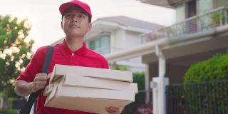亚洲送货男工人穿着红色制服搬运食物，披萨给年轻漂亮的女顾客在房前。邮差和快递杂货配送服务理念