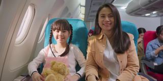 亚洲母女坐在飞机上靠近窗口，微笑着看着镜头。当飞机在天空中飞行时，女孩抱着熊娃娃。航空运输的概念。