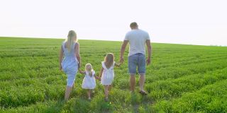 一个年轻快乐的四口之家带着两个孩子在绿地上散步。一家人带着孩子，孩子们在夏天的田野上散步。母亲，父亲和两个小女儿一起在户外享受大自然。美丽、健康、爱情、旅游、夏日时光的概念