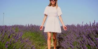 生活方式与一个年轻的金发白种女人漫步在一个夏天的下午紫色的薰衣草田在白色的裙子和帽子。在薰衣草花丛中漫步