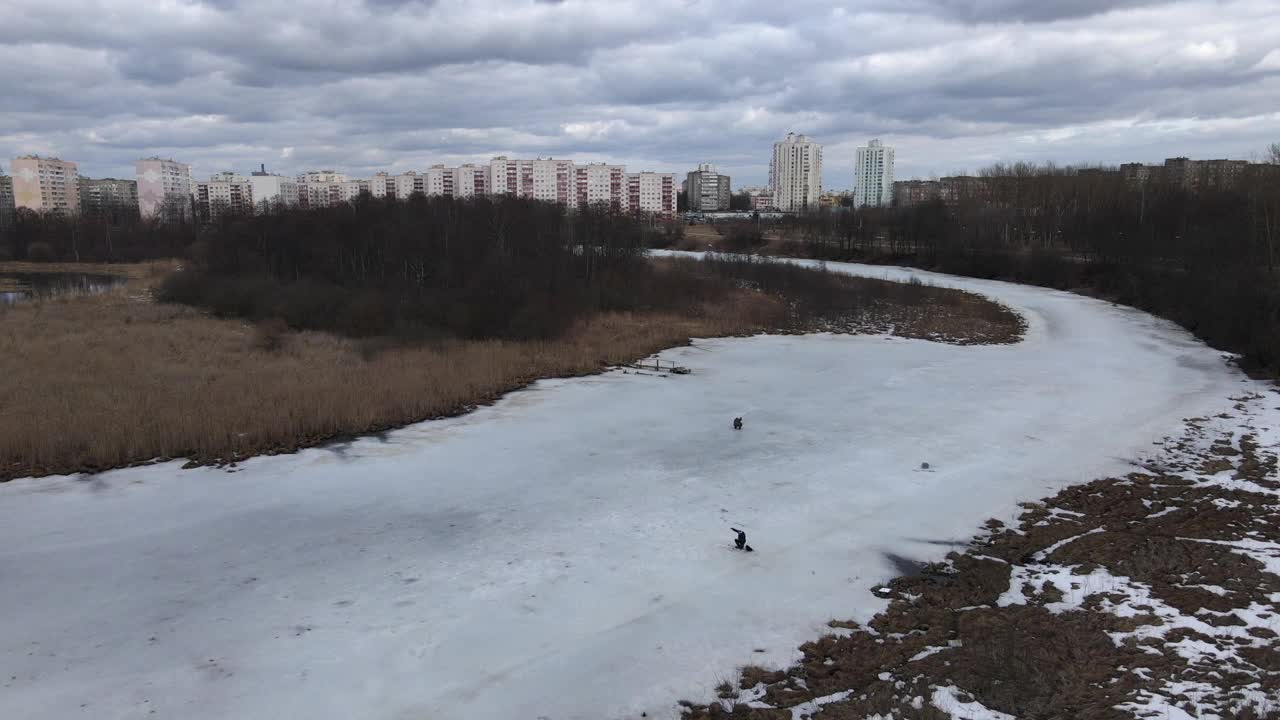 飞过一条结冰的河。渔夫们坐在冰上。冬季钓鱼。在地平线上可以看到多层建筑