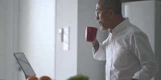 胡子花白头发的亚洲华人老人在厨房柜台边喝着咖啡边用着平板电脑