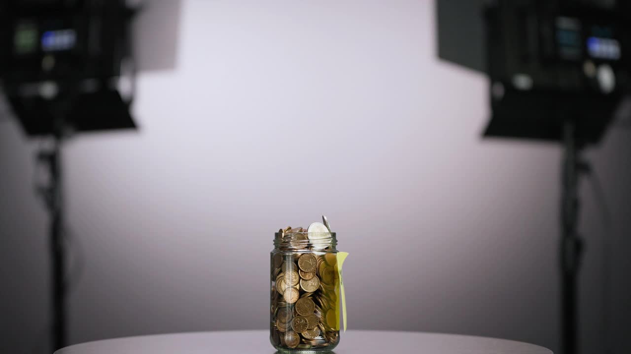 在后台拍摄的一个旋转玻璃罐，里面装满了硬币和两枚欧元硬币，上面贴着带有加密文字的粘性纸条，灰色背景，慢镜头