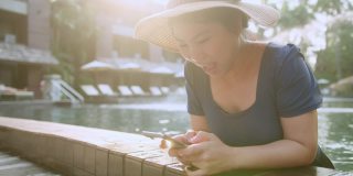 亚洲女性旅行者穿着休闲服装，享受假期日落时刻手用智能手机设备社交媒体交流。亚洲女性享受周末旅游游泳池日落时间。