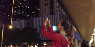 香港，一名年轻女子在晚上用手机拍摄建筑物。